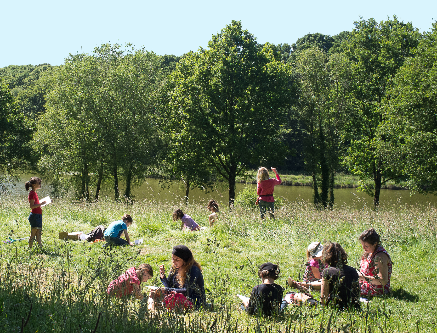 Adultes et enfants assis dans l'herbe, qui dessinent près d'un étang